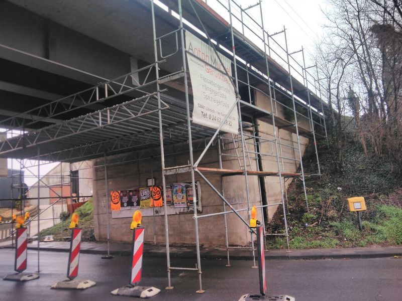 Gerüst unter einem Brückenüberbau in einer Unterführung, die Arbeitsebene reicht über die gesamte Breite, davor Leitbaken.