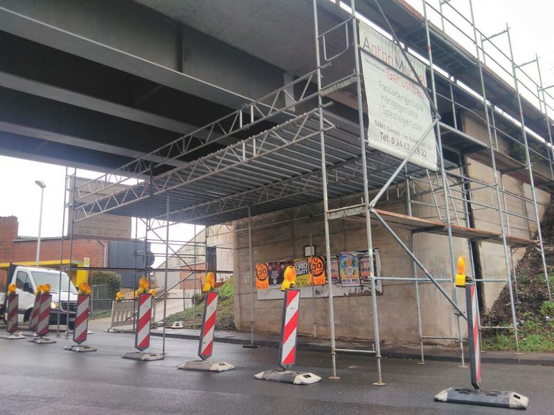 Abgehängte Arbeitsebene eines Gerüsts unter einem Brückenüberbau, auf der Fahrbahn davor stehen rot-weiße Leitbaken.