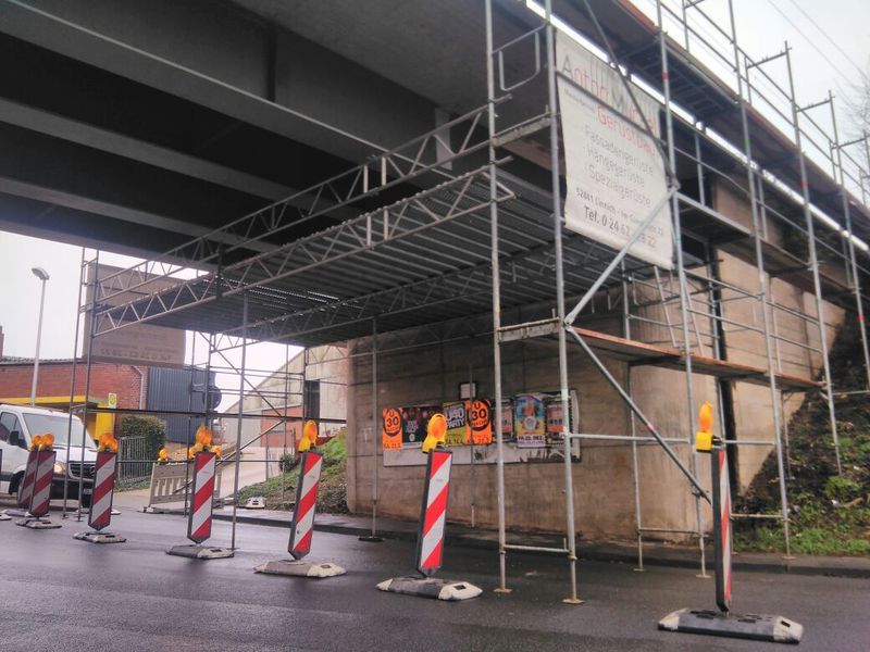 Seitlich abgefangenes Gerüst am Widerlager einer Brücke, auf der nassen Fahrbahn stehen Leitbaken mit gelben Warnleuchten.